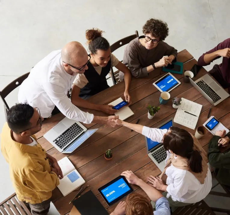 Overhead view of a team meeting around a conference table, two colleagues shaking hands over laptops and tablets